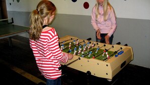Dos personas jugando al fútbol de mesa en una habitación con piso de madera y una pared decorada con patrones circulares coloridos.