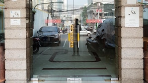 Entrada de un edificio con puerta de vidrio, un cartel que indica zona de estacionamiento y vista de vehículos estacionados y edificios al fondo.
