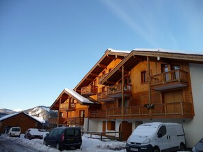 Eine Holzhütte mit mehreren Balkonen, einem überdachten Parkplatz und einem verschneiten Berg im Hintergrund.
