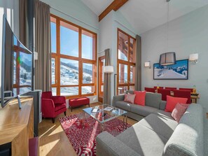 A modern living room with a sofa, red chairs, a glass coffee table, and large windows with a snowy mountain view.