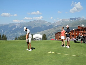 Ein Golfplatz mit Bergkulisse, eine Lodge mit roter Fassade und klarer Himmel.