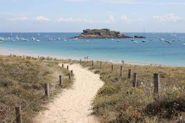 A sandy beach path leading to a rocky island with boats in the water.