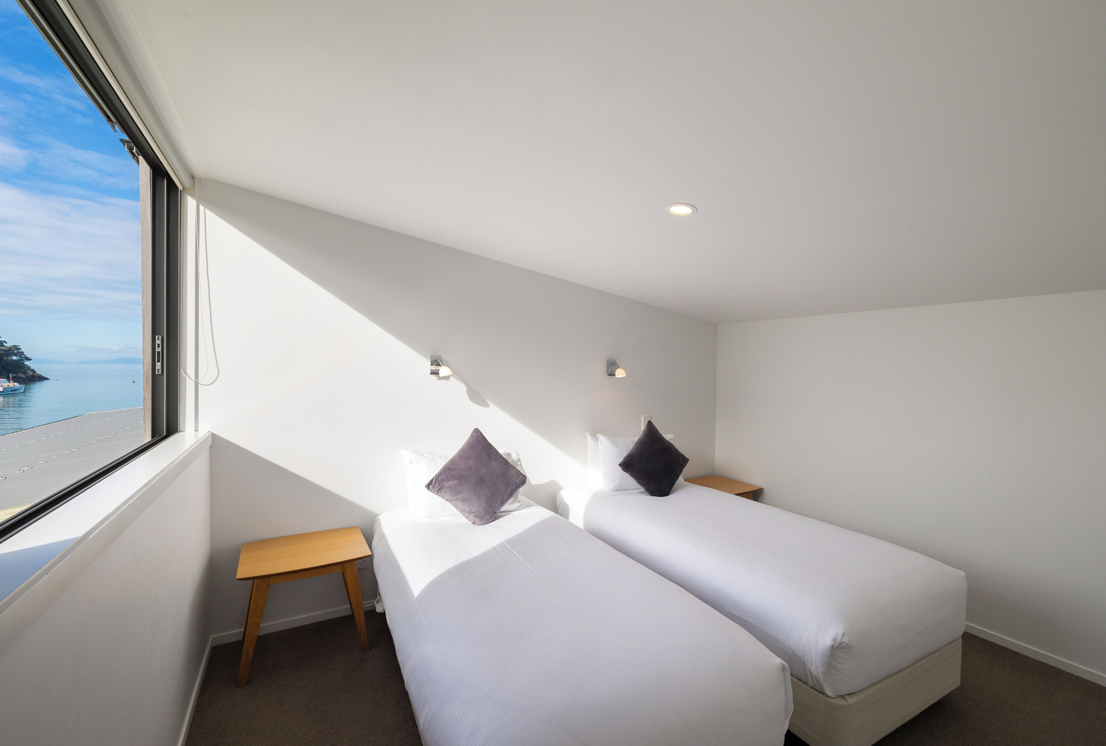 A room with two beds, a small wooden bench, a window with a view of the sea, and a ceiling light.