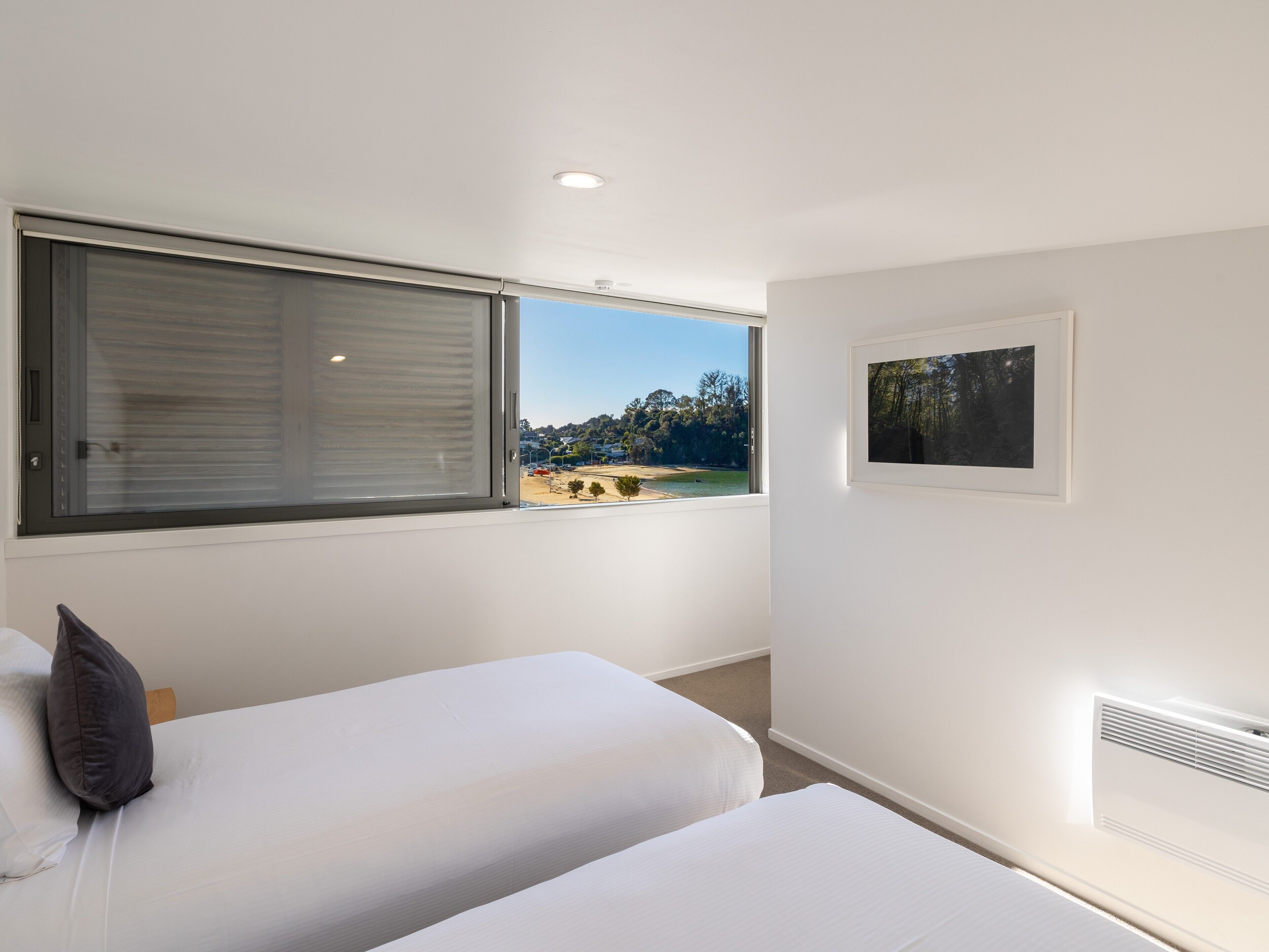 A bedroom with two beds, a window with blinds, a framed picture on the wall, and a view of a beach and trees.