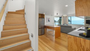 A modern interior with a wooden staircase, a kitchen with a stainless steel refrigerator, a grey sofa, and a large window overlooking a body of water. - Kaiteriteri Reserve Apartments (Kaiteriteri)