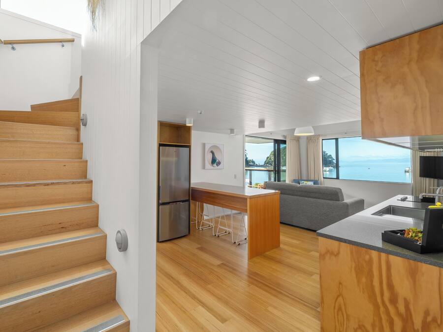 A modern interior with a wooden staircase, a kitchen with a stainless steel refrigerator, a grey sofa, and a large window overlooking a body of water.