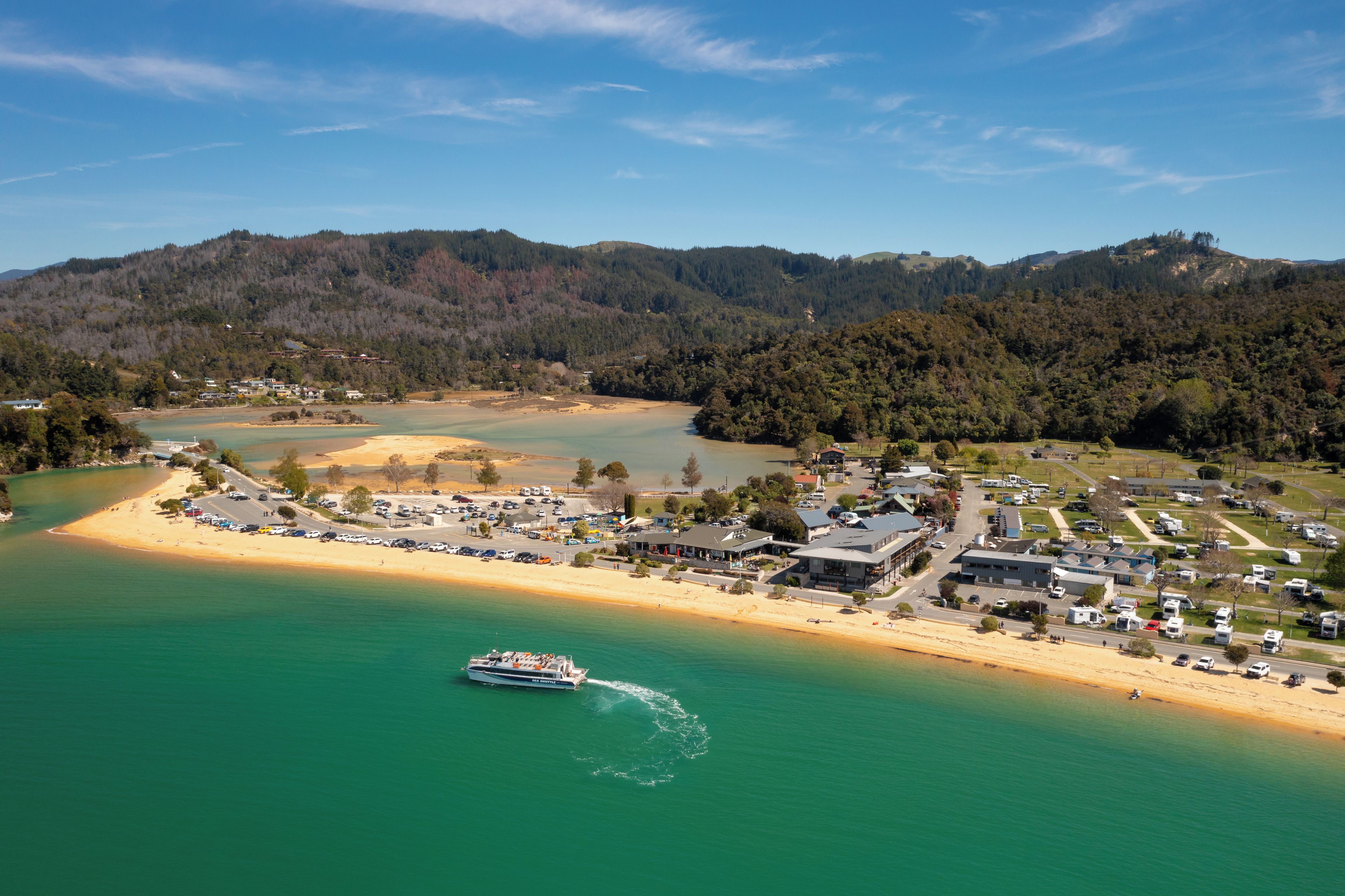 Une ville côtière dotée d’une plage, d’un bateau-navette maritime et d’un paysage vallonné en arrière-plan.
