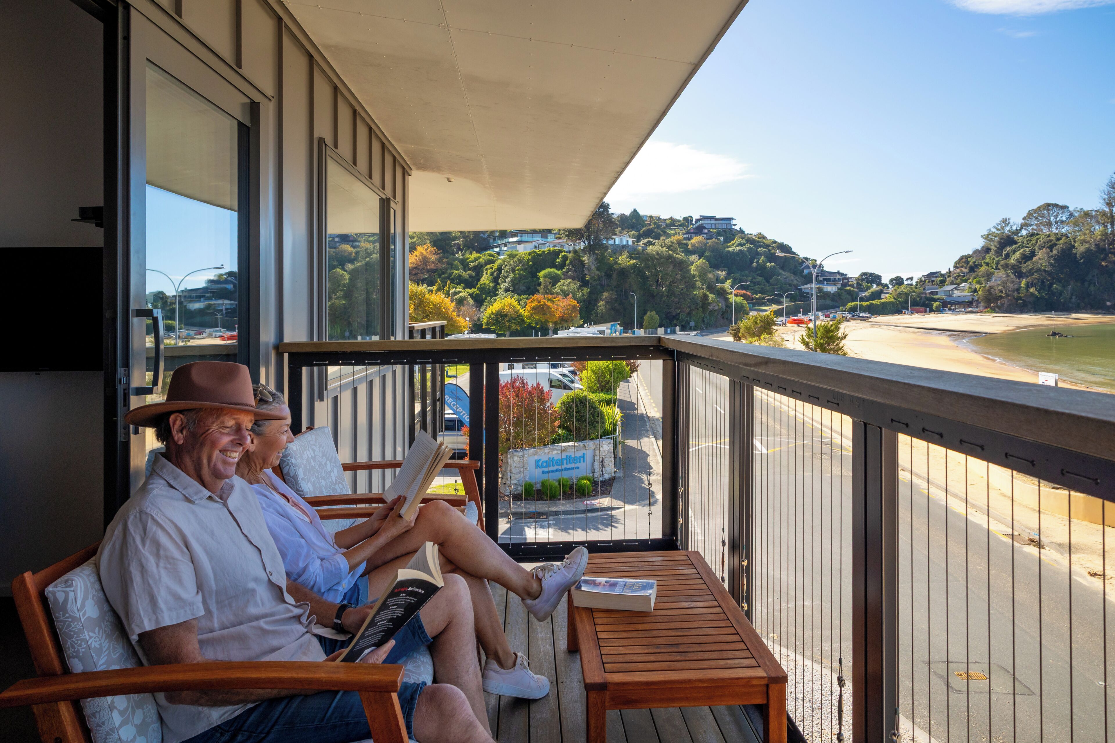A balcony with two people reading, a wooden table, and a view of a beach and buildings.