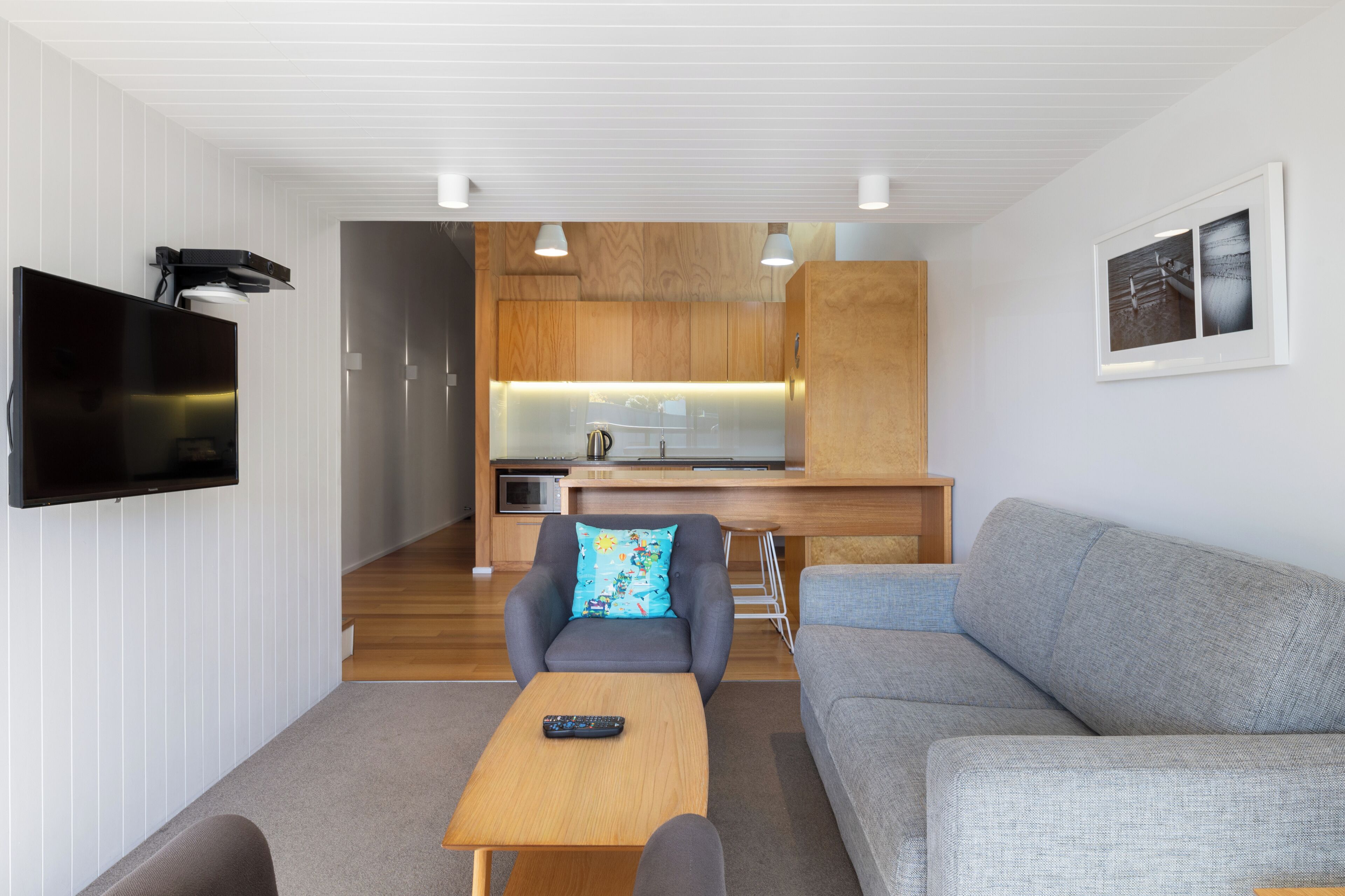 A modern living room with a flat-screen TV, a grey sofa, a wooden coffee table, and a kitchen in the background.