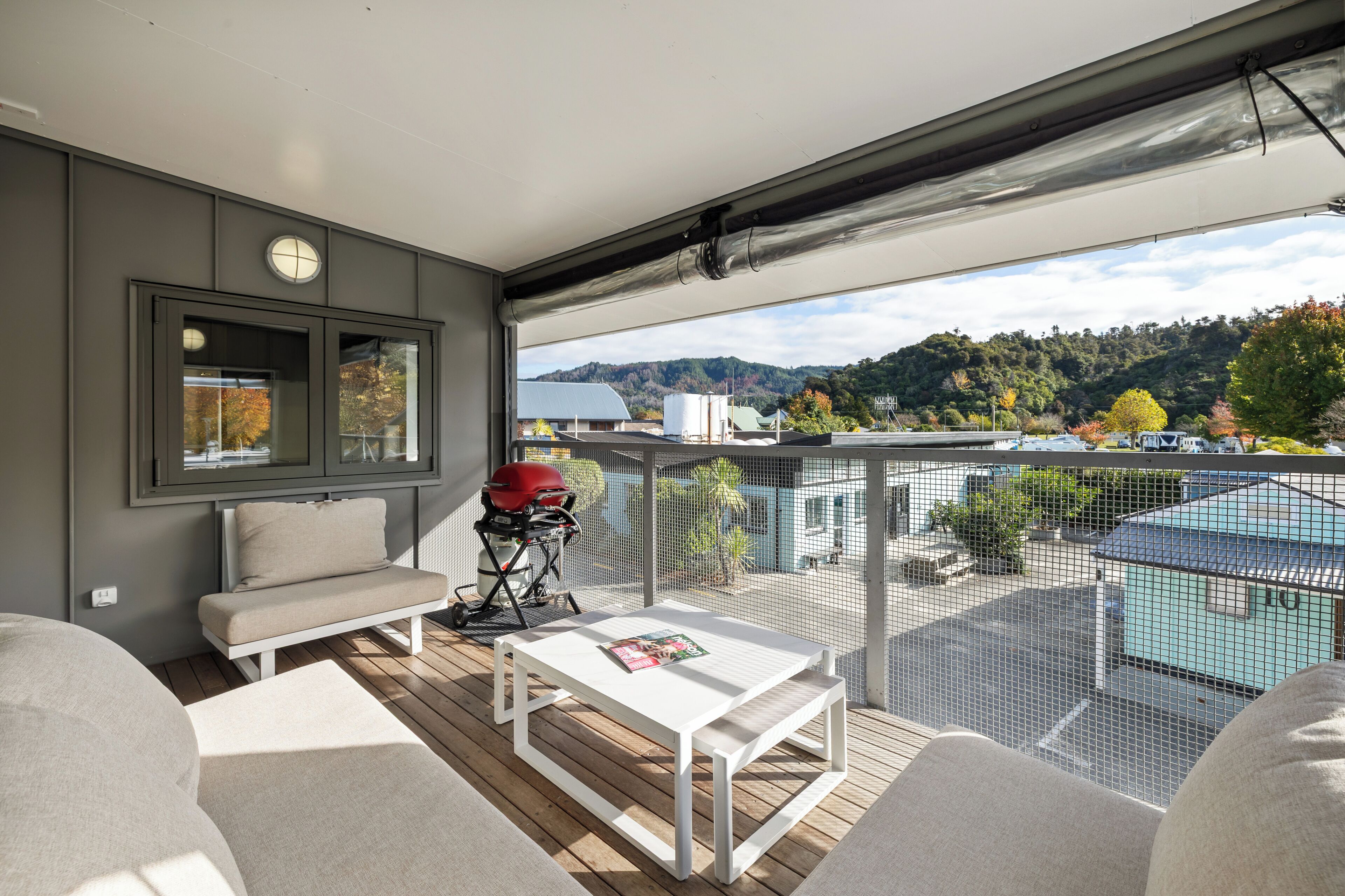 A balcony with a white table, a red grill, and a view of a mountainous landscape.