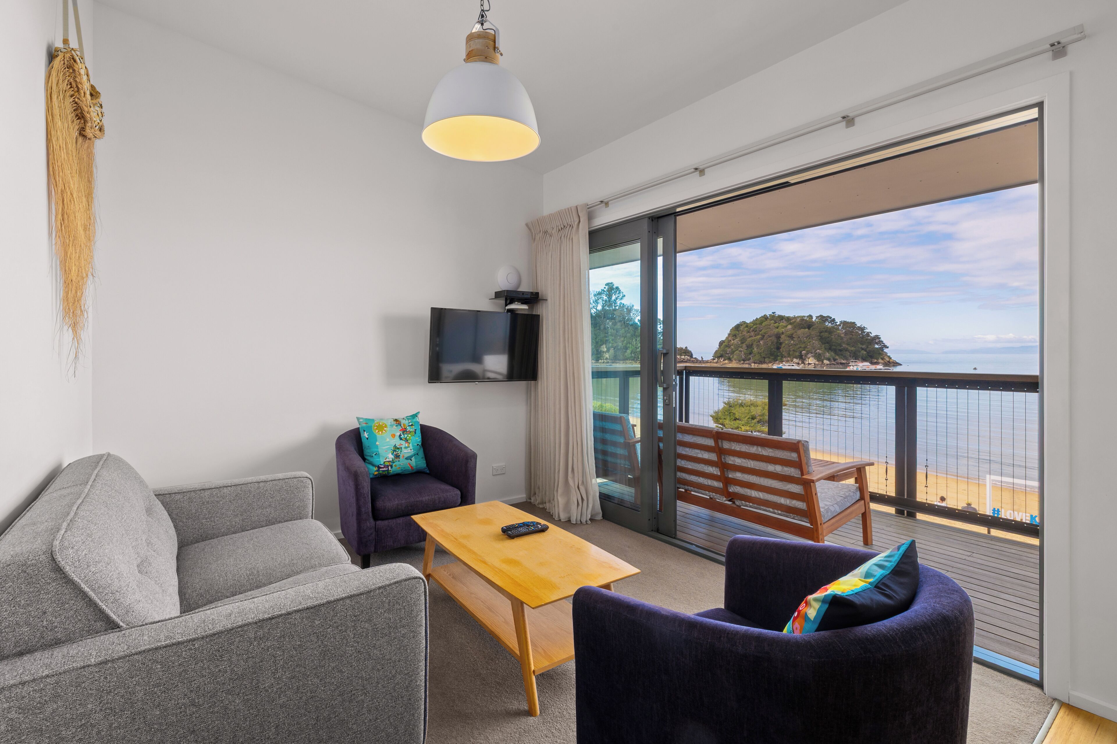 A modern room with a sofa, armchair, and a table, a TV mounted on the wall, and a balcony with a view of the sea.