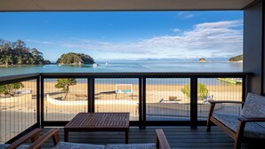 A balcony with a view of a beach and water, wooden furniture, and a signboard. - Kaiteriteri Reserve Apartments (Kaiteriteri)