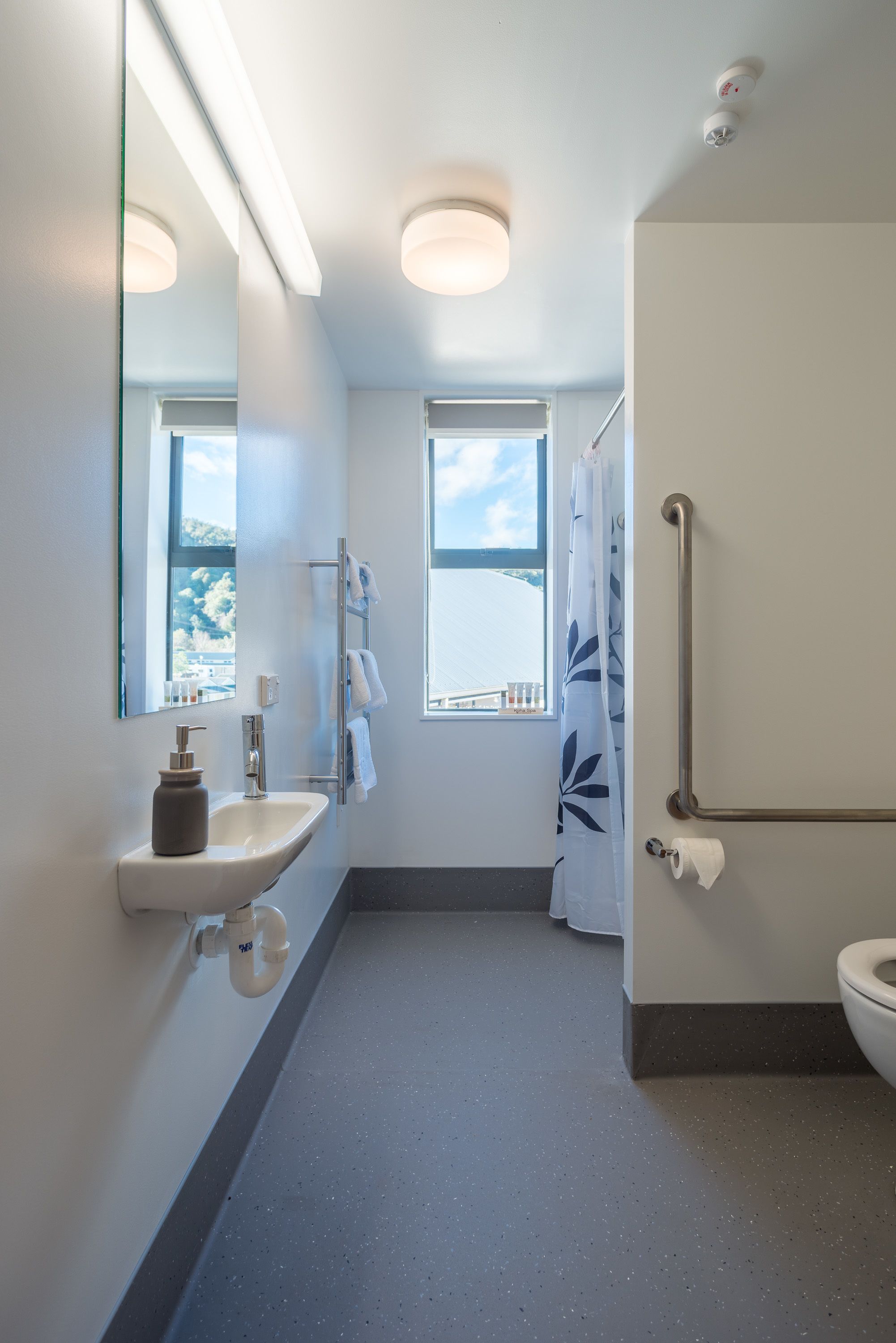 A modern bathroom with a sink, mirror, and toilet. It features a window with a view of the sea and a shower area with a grab bar.