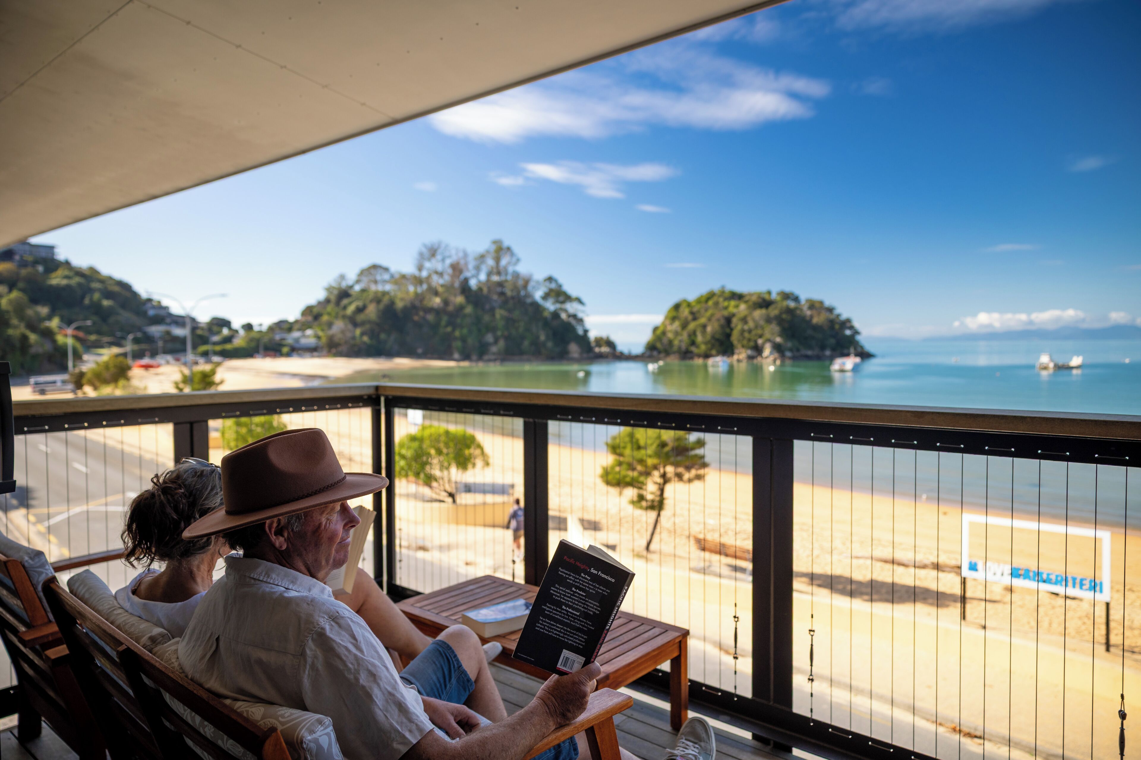 Un couple est assis sur un banc en bois, face à une plage, d’où l’on a une vue dégagée sur l’océan et les îles lointaines.