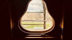 A window with a unique pear-shaped frame overlooking a grassy field with trees. - Cozy Forest Chalet Escape (Waimes - Sourbrodt)