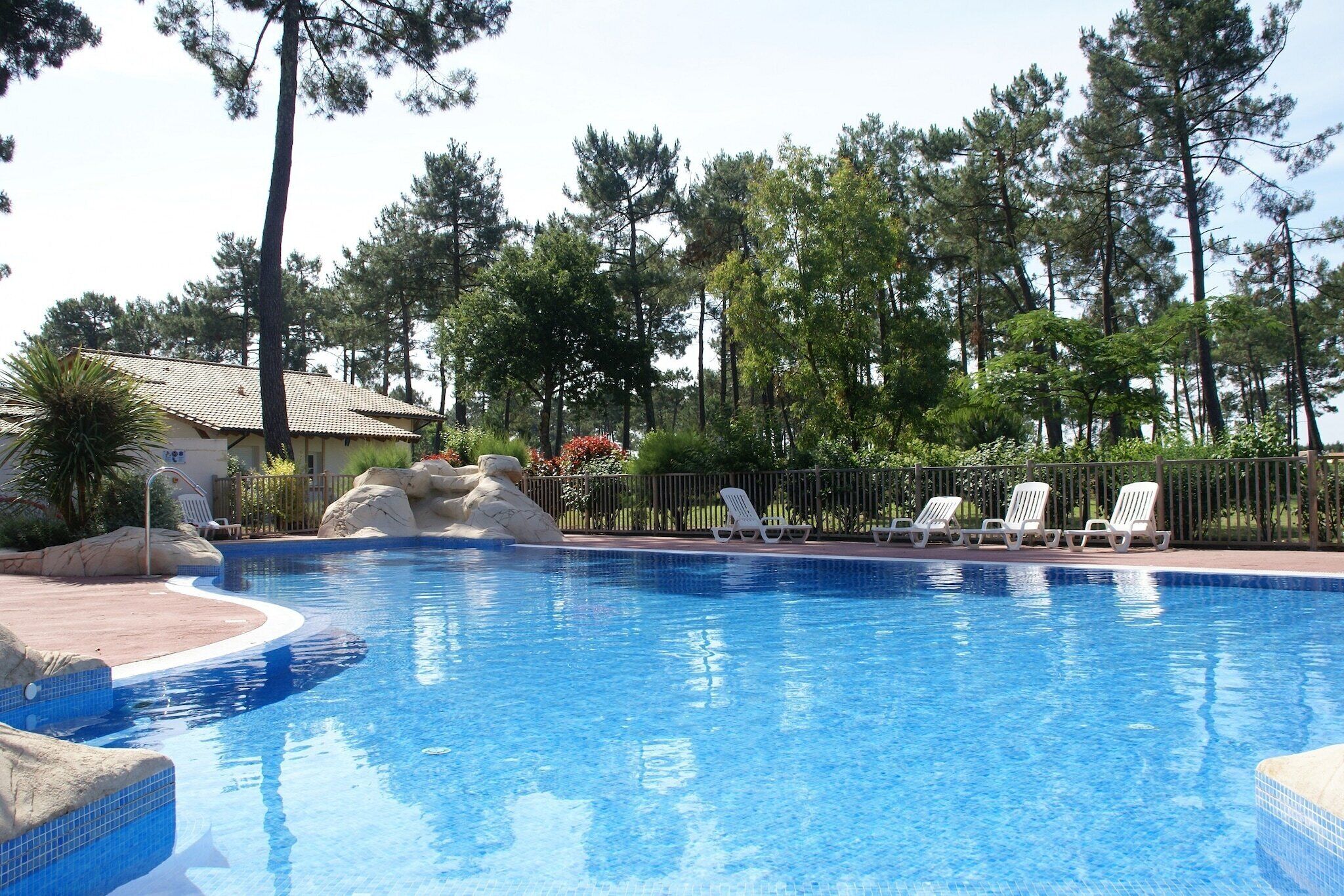 A swimming pool with a tiled edge, surrounded by a fence, and white lounge chairs.