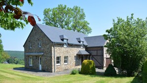 Une maison en pierre avec un toit en pente, une pelouse bien entretenue et un grand arbre à l’avant.