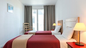 Hotellihuone, jossa on kaksi sänkyä, tuoli, yöpöytä lampun kanssa ja ikkunasta avautuva näkymä ulos.
