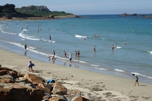 A beach with people swimming and walking along the shore, with rocky outcrops in the foreground.