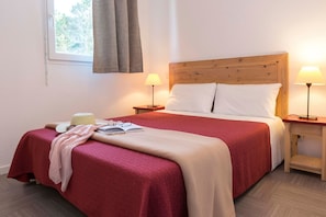 A neatly made bed with a red and beige bedspread, two bedside lamps, a window with curtains, and a small bedside table with a lamp.
