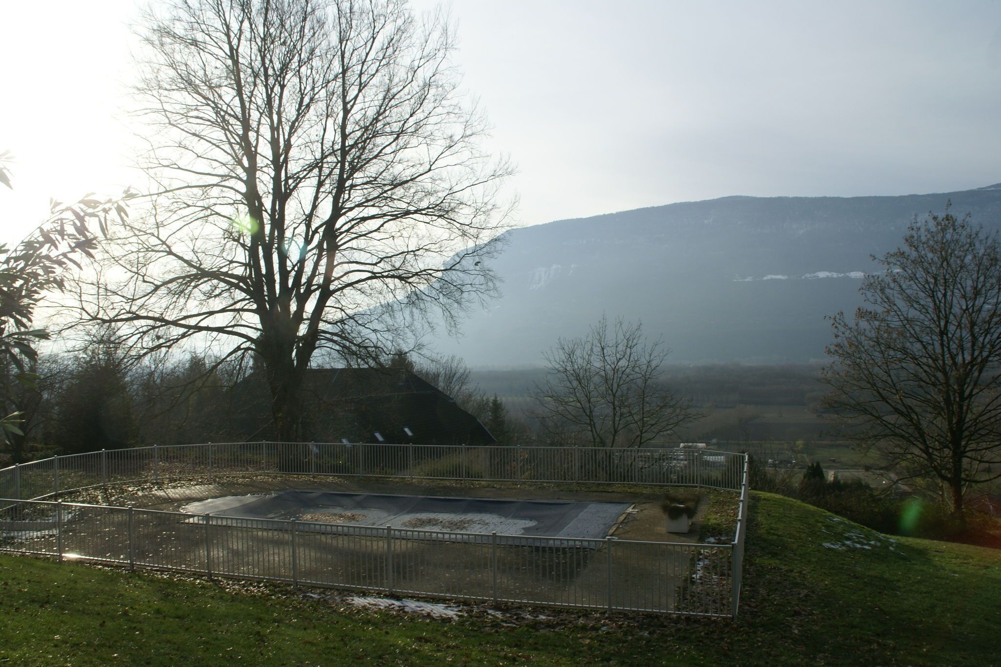 A fenced outdoor area with a metal structure, possibly a pool or water feature, surrounded by trees and a hilly landscape in the background.