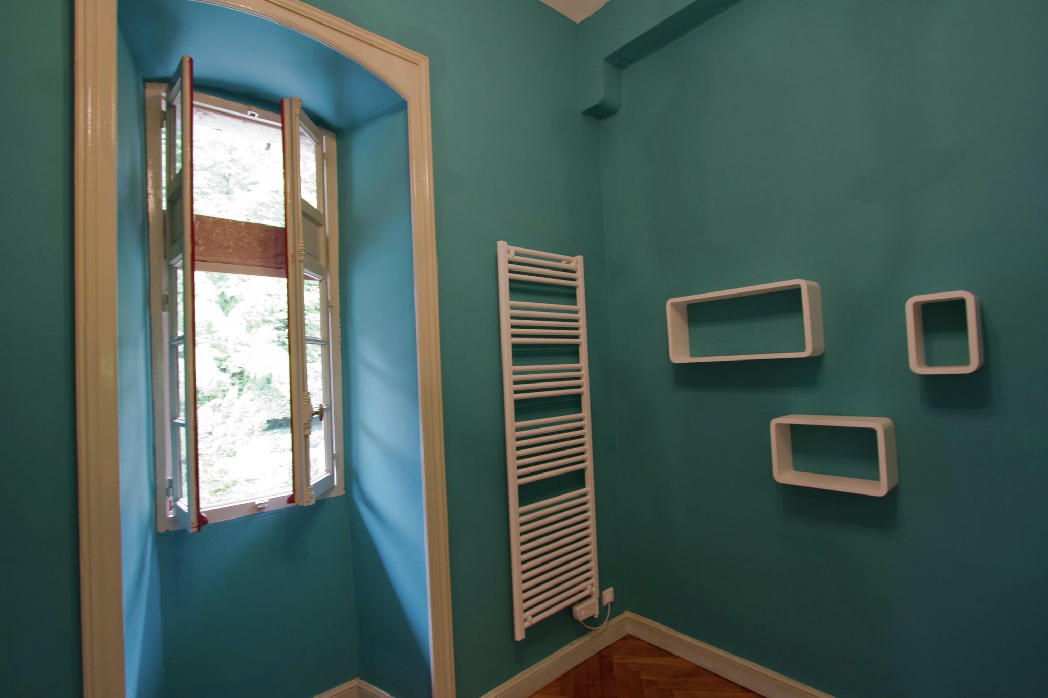 A room with teal walls, a window with white trim, a white radiator, and three white wall-mounted shelves.
