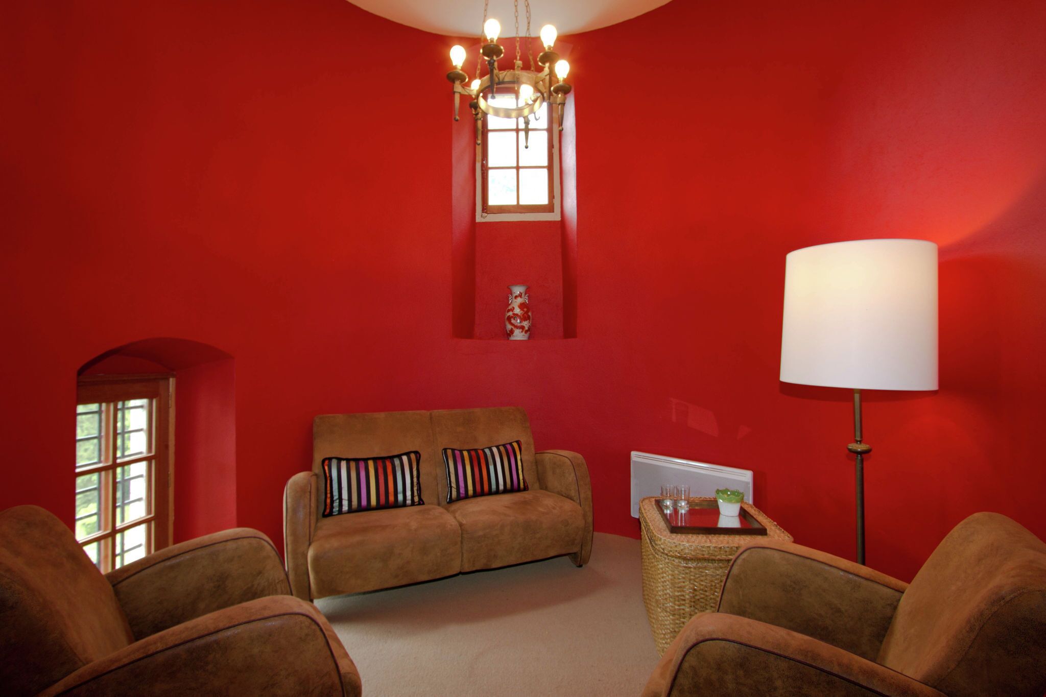 Una habitación con una pared roja, un sofá marrón con almohadas a rayas, una lámpara de pie y una ventana con vistas a la vegetación.