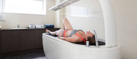 A person lying in a modern, white, freestanding bathtub in a bathroom with a window, a sink, and a towel on the counter.
