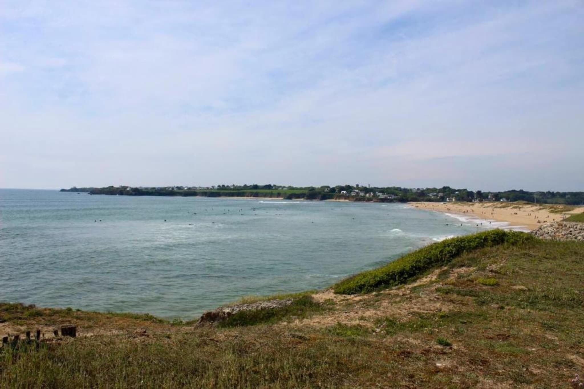 Un paysage côtier avec une plage, l’océan et une côte lointaine parsemée de bâtiments.