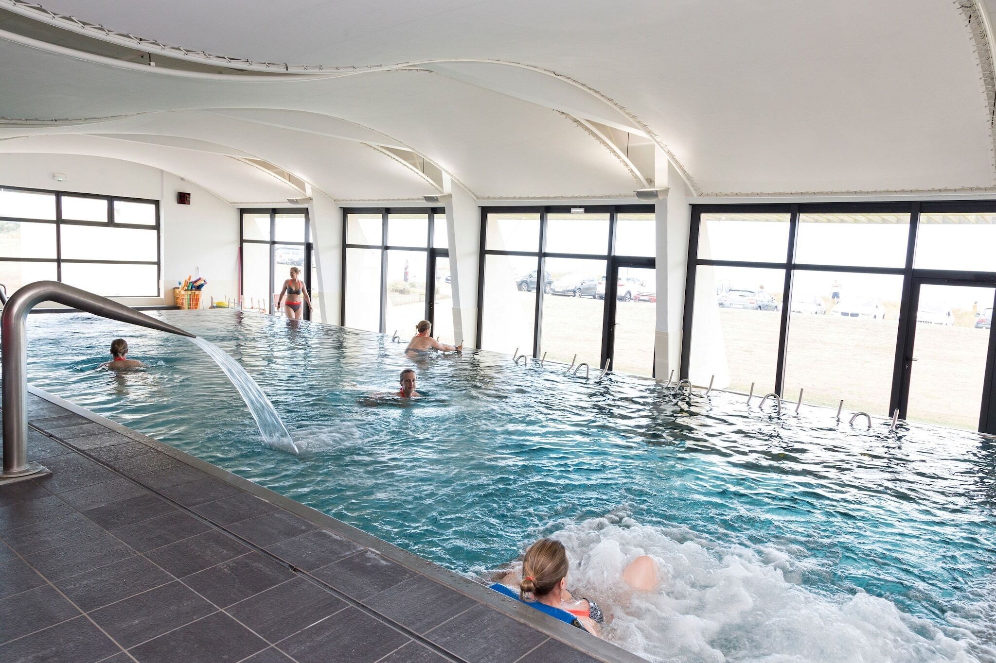 Uma piscina interna com janelas transparentes, chão revestido e uma pessoa na água.