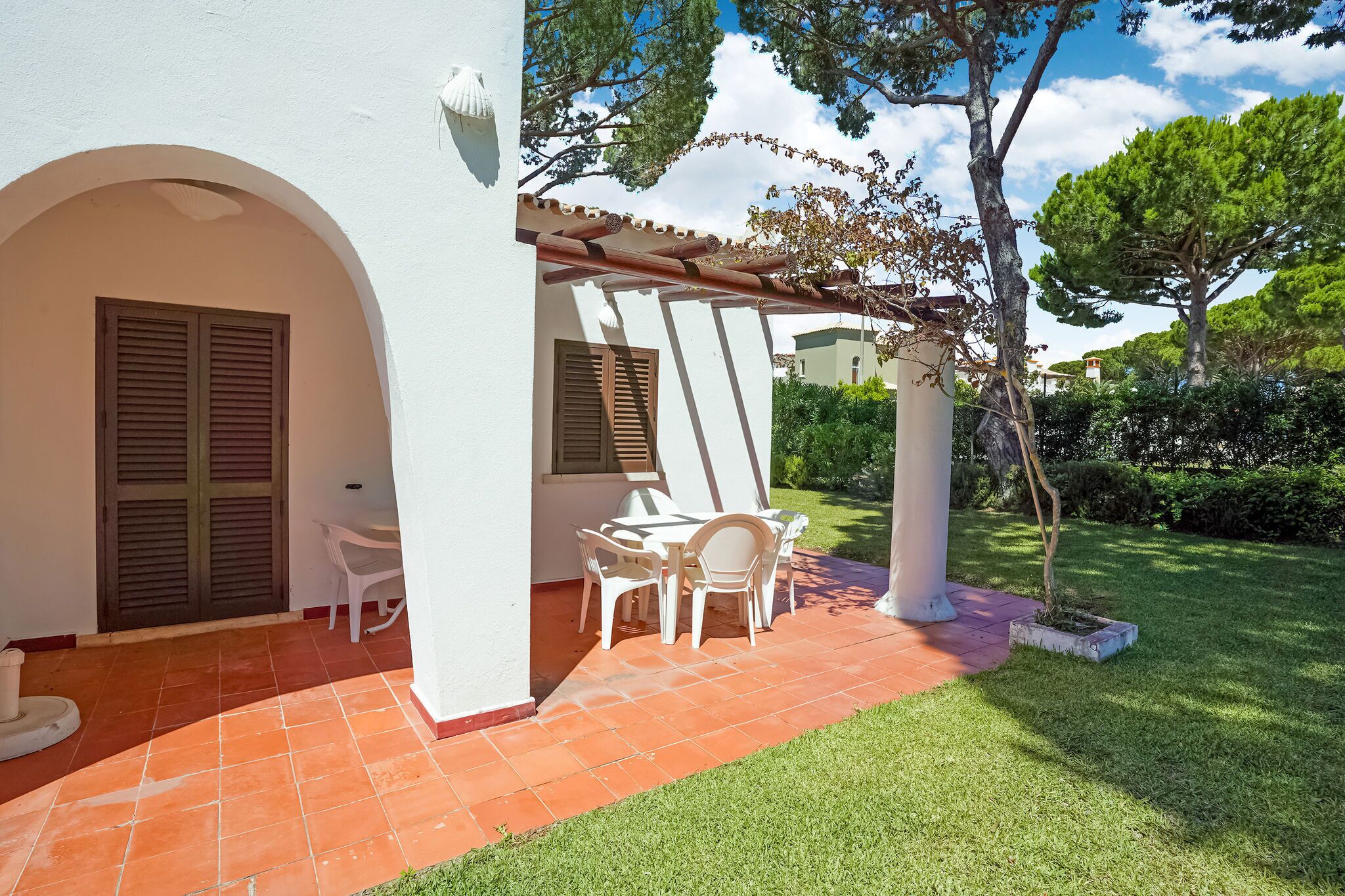 A white building with a tiled patio, a pergola, and a small garden area with a table and chairs.