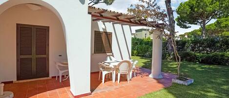 A white building with a tiled patio, a pergola, and a small garden area with a table and chairs.