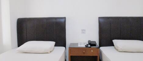 A room with two beds, each with a pillow and a red blanket. There is a small wooden nightstand between the beds with a phone and a lamp.