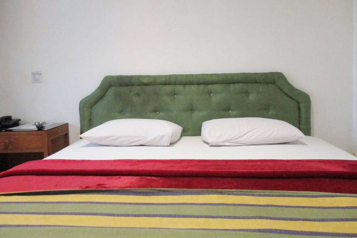 Un lit avec une tête de lit recouverte d’un tissu vert, deux oreillers blancs et une couverture à rayures rouges et jaunes.