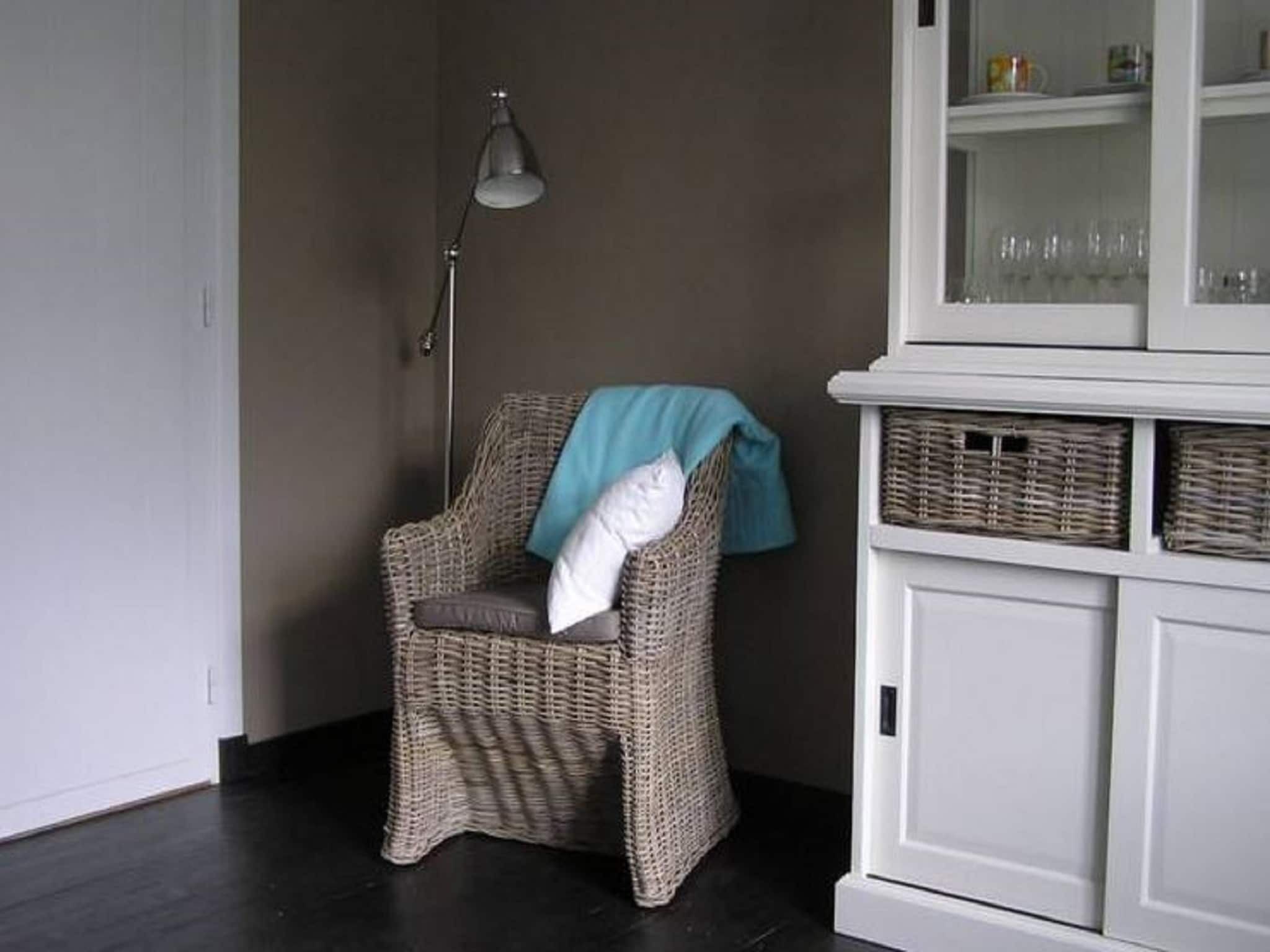 Een rieten stoel met een wit kussen en een blauwe handdoek, een vloerlamp en een witte kast met manden.
