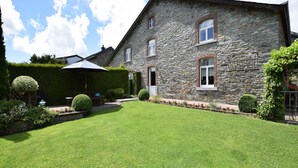 Une maison en pierre avec une pelouse bien entretenue, une terrasse avec une table et des chaises, et une haie.