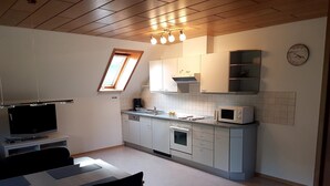 Cocina funcional con microondas, horno y lavabo. Hay una mesa de comedor con sillas y un televisor sobre un mueble. Un tragaluz se encuentra sobre el lavabo y un reloj en la pared.