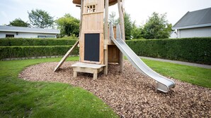 Een houten speelconstructie met een glijbaan en klimelementen, omringd door een verzorgd gazon en heggen.
