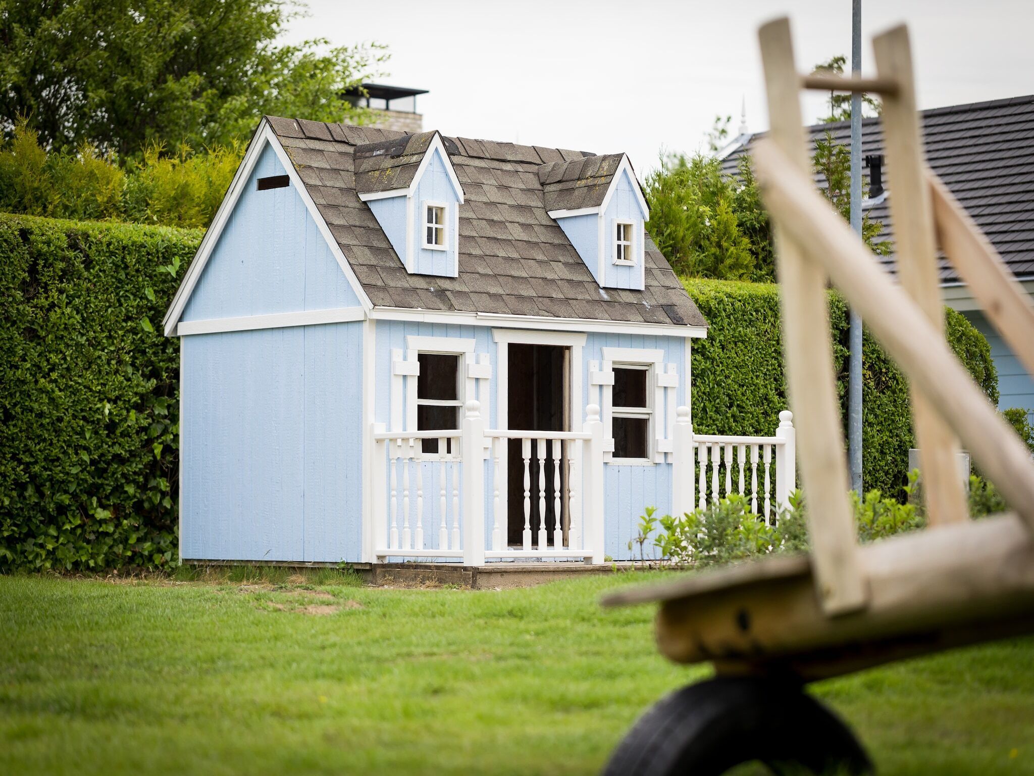 A small, light blue wooden house with a gabled roof and white trim, surrounded by a well-maintained hedge and grass.