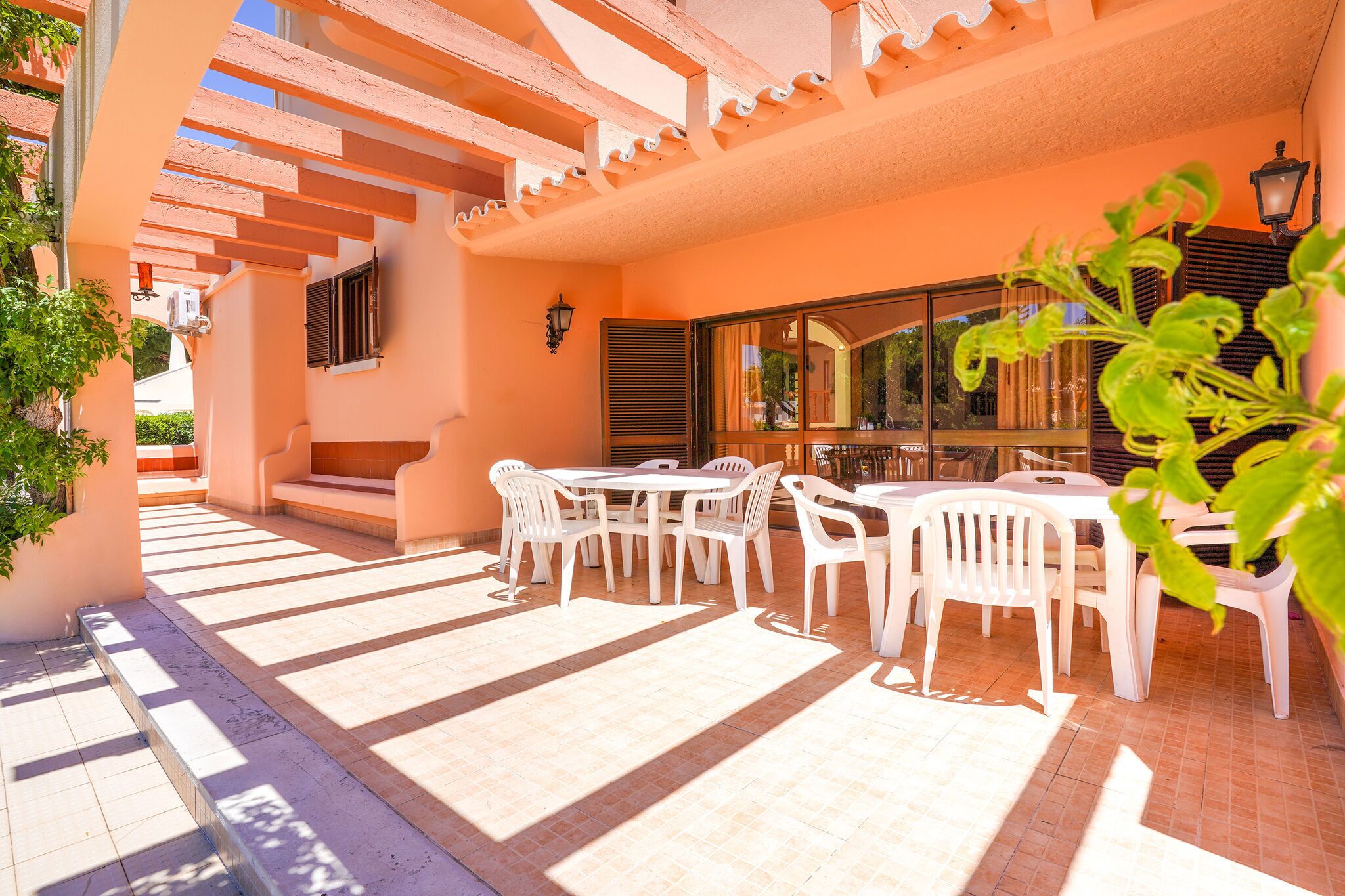 Un patio al aire libre con sillas y mesas de plástico blanco, una pérgola de madera y un ventanal con persianas.