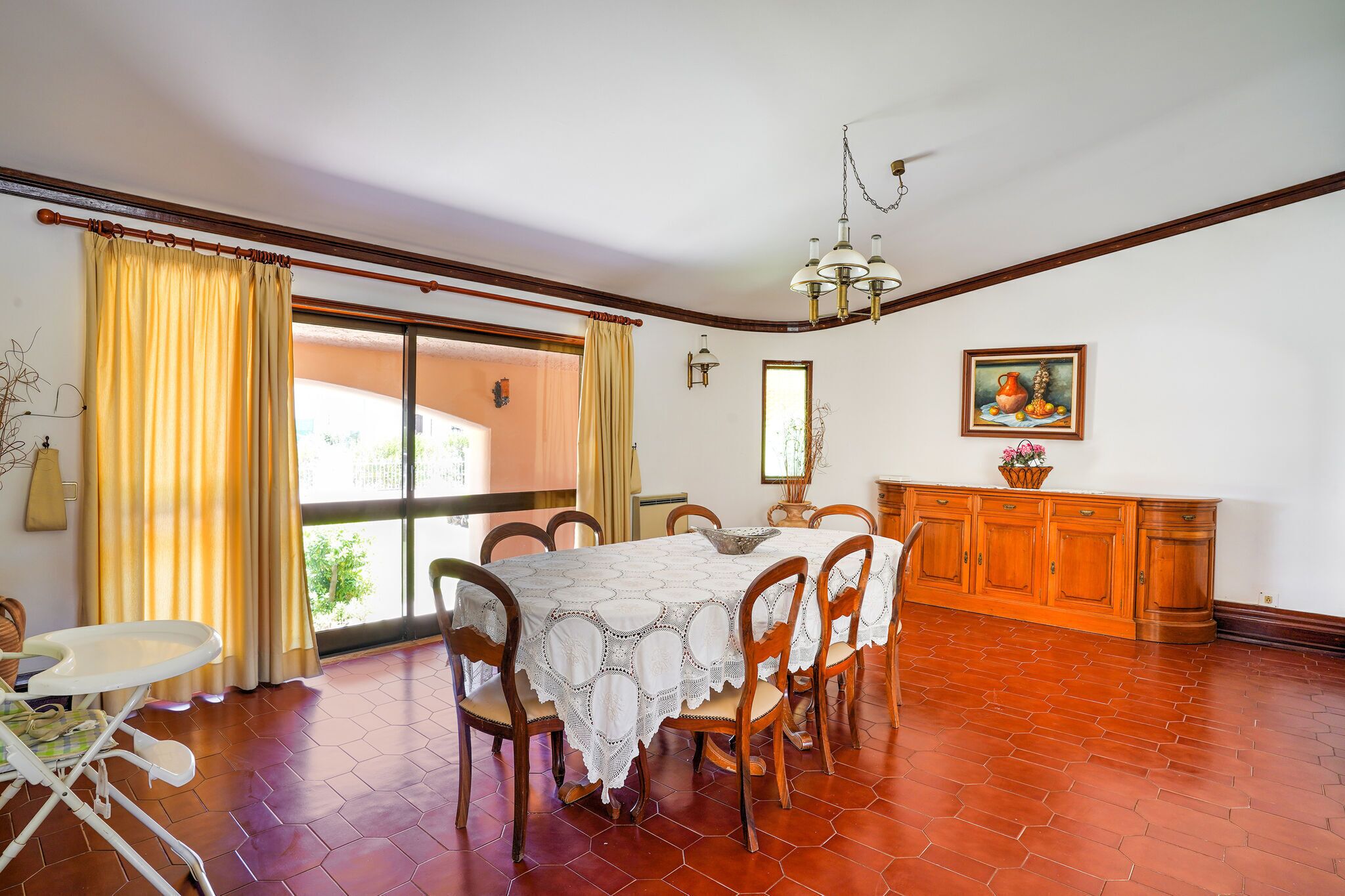 Un comedor con una mesa dispuesta para seis personas, sillas de madera, mantel blanco, una araña y vistas a través de un ventanal.