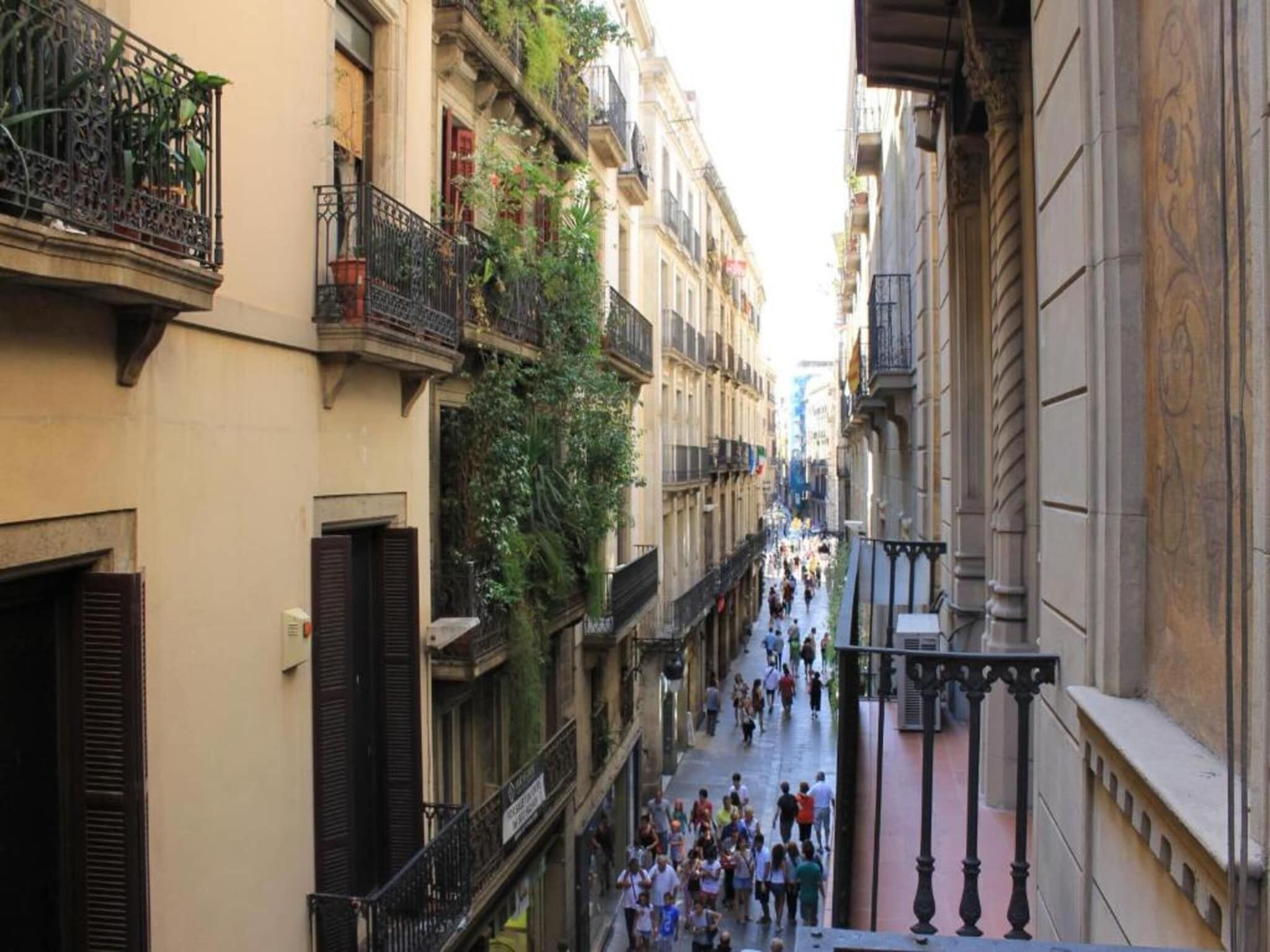 Une rue étroite en milieu urbain, bordée de bâtiments des deux côtés, avec des balcons et des piétons.