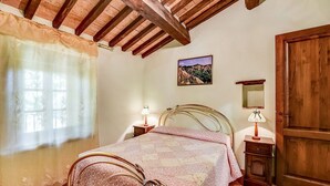 A bedroom with a bed, wooden ceiling beams, a window with curtains, a bedside table with a lamp, and a door. - Belvilla by OYO Sorgente (Radicofani)