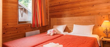 Une chambre dans un chalet en bois, avec deux lits, une fenêtre avec des rideaux, une lampe et une petite table.