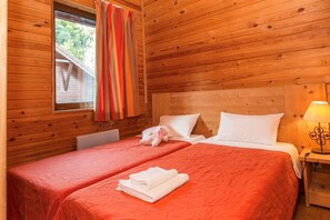 Ein Zimmer in einer Holzhütte mit zwei Betten, einem Vorhangfenster, einer Lampe und einem kleinen Tisch.