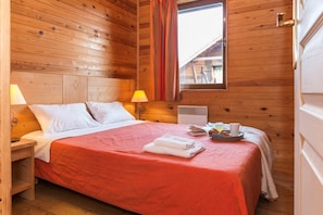 A wooden room with a bed, a nightstand, a lamp, a window with curtains, and a tray with breakfast items.