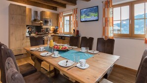 Un coin repas avec une table en bois dressée pour six personnes, une cuisine en arrière-plan et une vue pittoresque par la fenêtre.