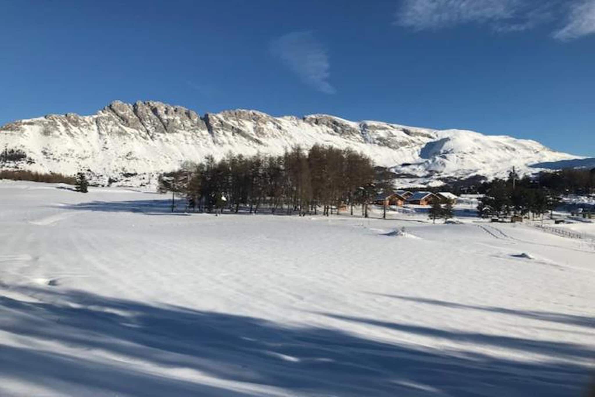 A snow-covered landscape with a mountain range, a cluster of trees, and a few buildings in the distance.