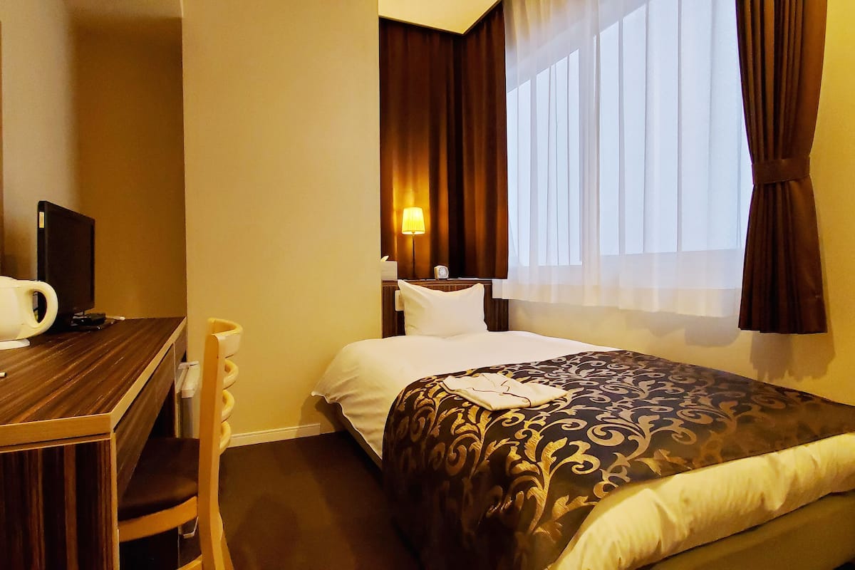 A hotel room with a bed, a desk, a chair, a lamp, and a window with curtains.