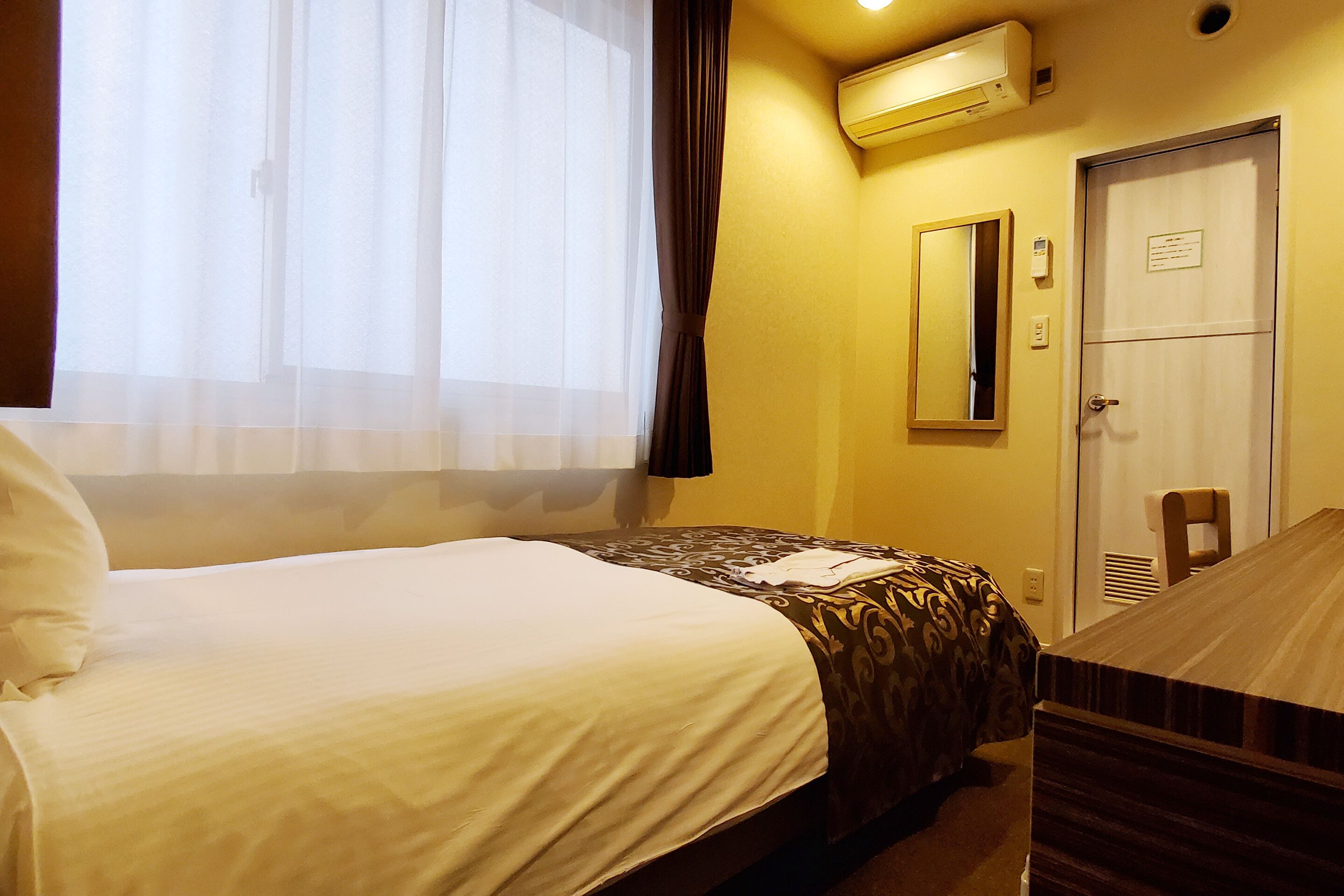 A hotel room with a bed, a desk, a mirror, and an air conditioning unit.